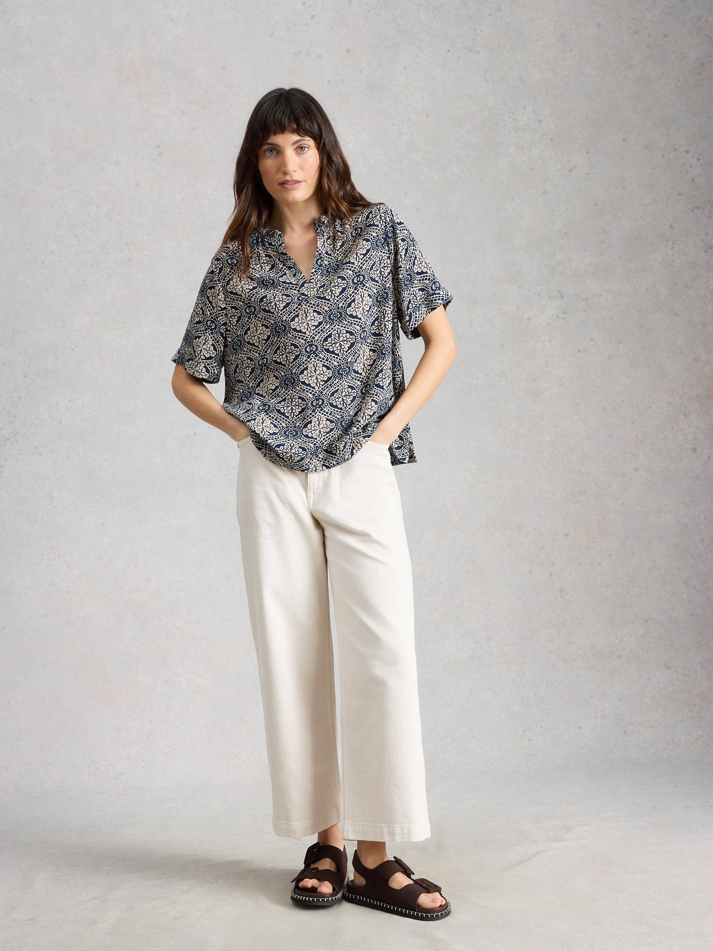 Woman wearing a patterned blouse and white pants against a plain background