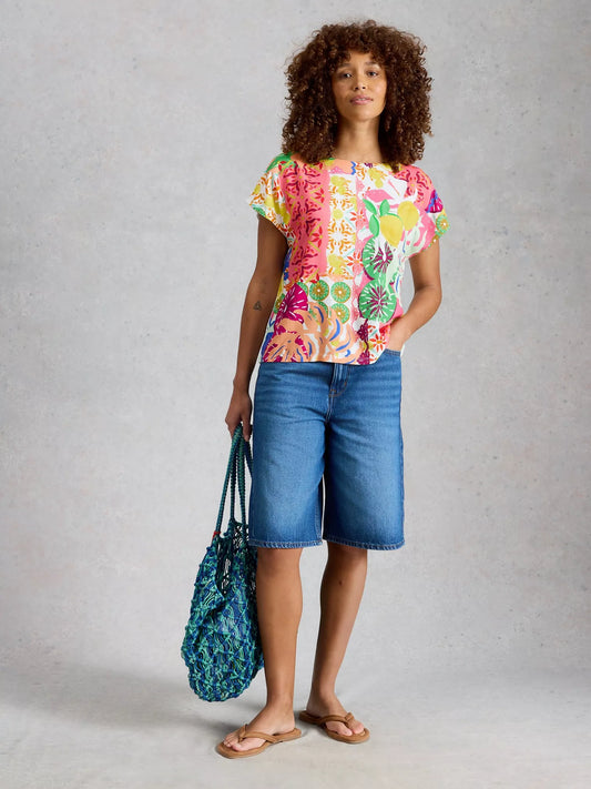 Woman wearing a colorful floral top and blue shorts, holding a blue bag on a gray background