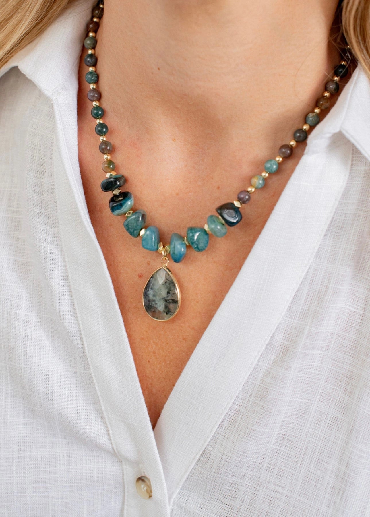 Necklace with gemstones and a teardrop pendant worn by a person in a white shirt.