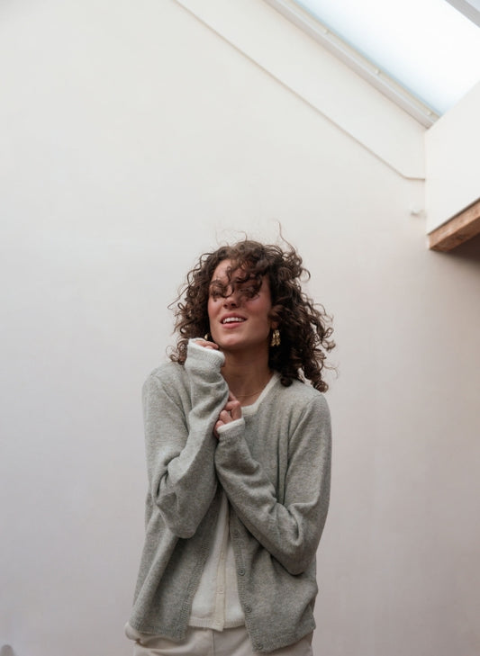 A woman with curly brown hair wearing a light gray knit cardigan over a cream top, standing against a minimalist off-white wall under a skylight.