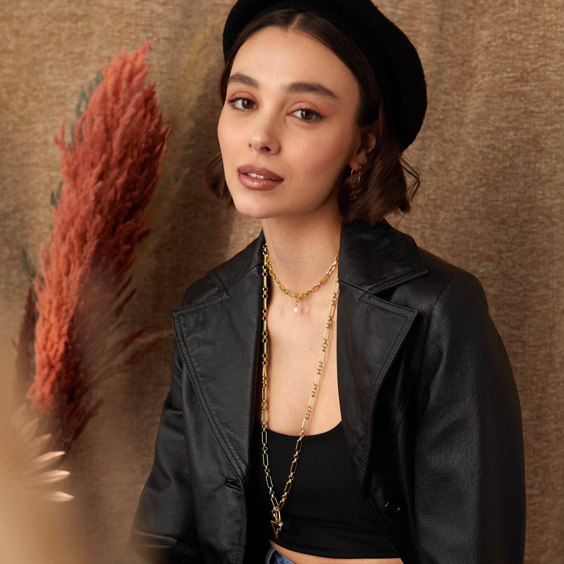 Woman wearing a black jacket and hat with gold necklaces against a brown background